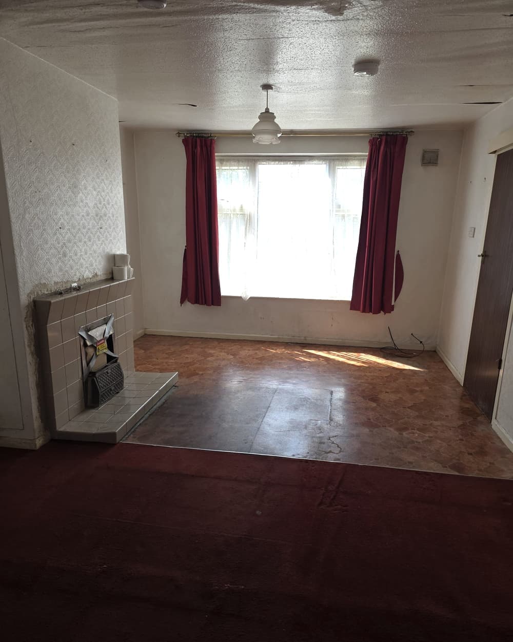 A cleared living room with red curtains, a tiled fireplace and sunlight on the floor
